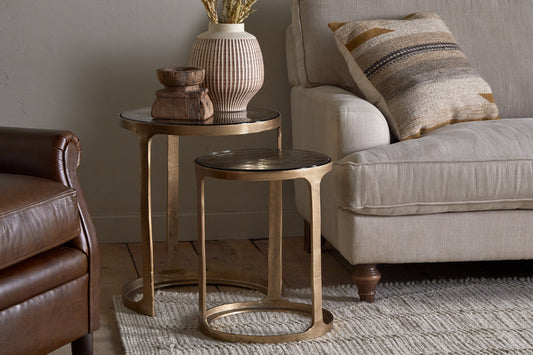 Pair of Nkuku Khidki cast metal and glass nesting side tables in antique brass finish, set of 2, with intricate metalwork and glass tops.
